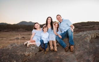 Family portrait outdoors with mountains in background, smiling and sitting on a rock.
