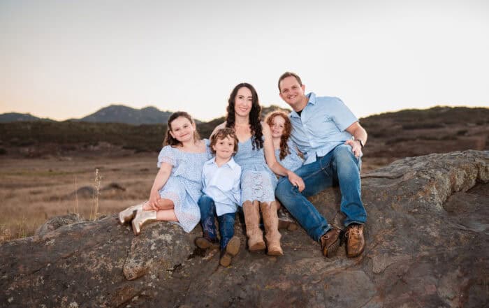 Family portrait outdoors with mountains in background, smiling and sitting on a rock.