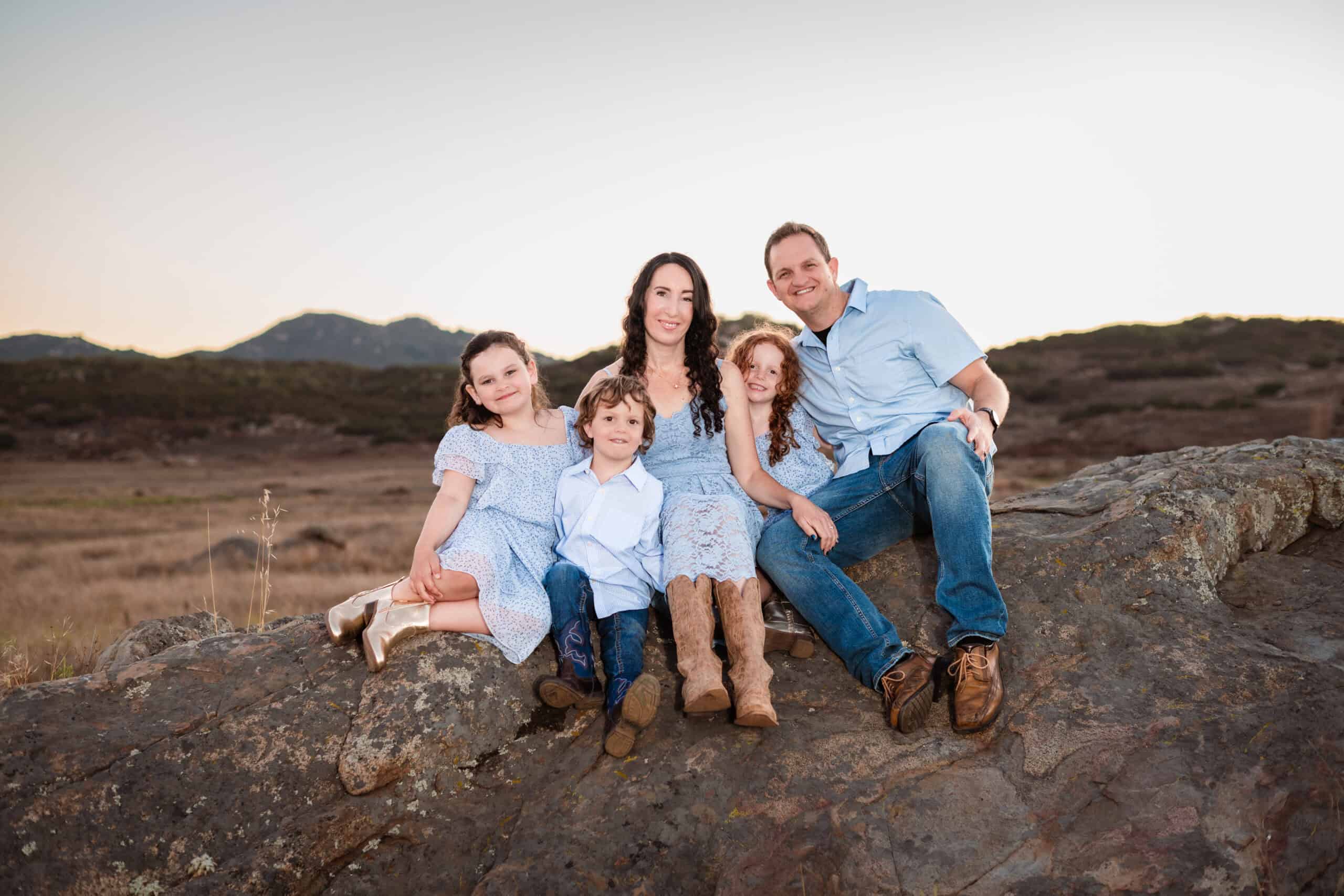 Family portrait outdoors with mountains in background, smiling and sitting on a rock.
