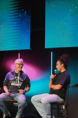 Two young women engaged in a conversation during a youth event at Canyon Springs Church, with vibrant stage lighting in the background. Youth discussion at Canyon Springs Church with two young women on stage.