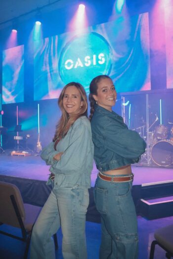 Two women smiling at a concert stage with Oasis logo in the background.